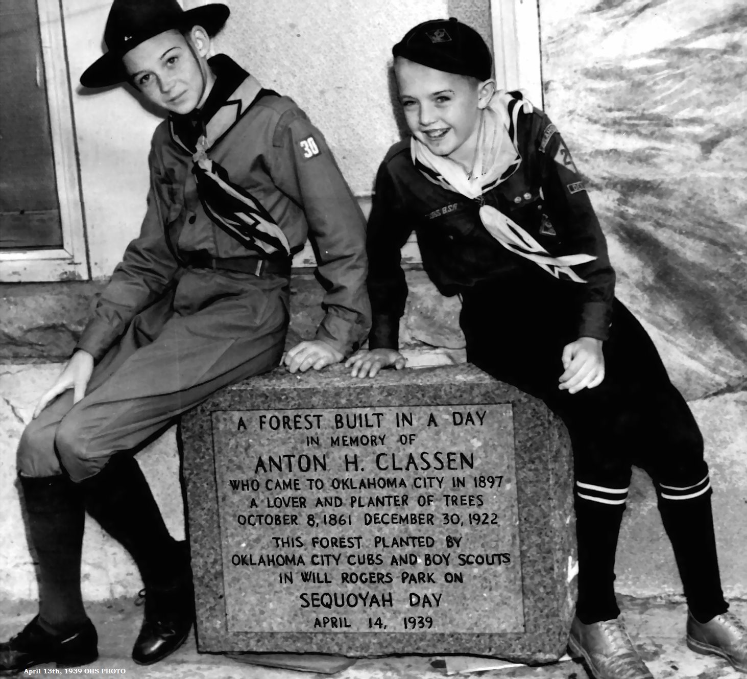 B/W picture of a boy scout and a cub scout sitting next to a plaque honoring